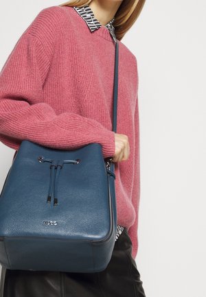 Vrouw draagt een roze gebreide trui en een blouse met patroon, en houdt een blauwe leren emmertas met schouderriem vast.