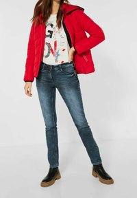 Chaqueta de plumas roja con capucha sobre una camiseta blanca con gráfico, combinada con jeans ajustados de color azul oscuro y botines negros de suela gruesa.