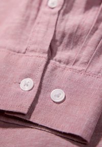 Camicia di lino rosa chiaro che presenta una patta con bottoni bianchi in plastica e una consistenza liscia. Con colletto e dettagli di cucitura intricati.