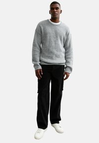 Gråstickad tröja med rund hals, matchad med svarta cargobyxor. Modellen bär vita sneakers. Tyget verkar vara strukturerat och varmt.