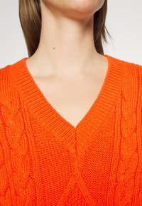 Personne portant un pull orange vif en maille torsadée à col en V, avec le cou visible et une partie du menton, devant un fond uni.