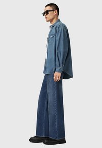 Ljusblå denimskjorta, oversized passform, med fickdetalj framtill. Mörkblå vida denimbyxor, slät textur, kombinerade med svarta solglasögon och skor.