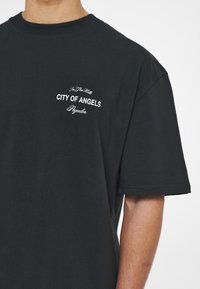 Černé tričko s krátkým rukávem vyrobené z bavlny, na kterém je bílý text: "In The Hills," "CITY OF ANGELS," a "Pagador." Jednoduchý design s kulatým výstřihem.