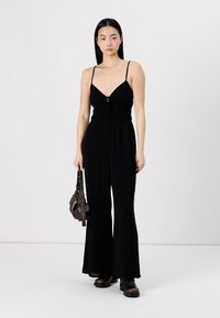 Macacão preto com corpete justo, alças finas ajustáveis e design de perna ampla. Estilizado com uma pequena bolsa castanha e botas.