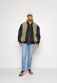 Olivinės žalios pūkinių liemenių virš juodo bombonešio tipo švarkelio, šviesiai mėlyni džinsai ir violetiniai-geltoni sportbačiai; neutralios spalvos šalikėlis; kasdienis stilius.