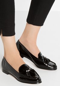 Des pieds chaussés de mocassins en cuir verni noir avec des pampilles, associés à un pantalon noir court sur un fond clair uni.