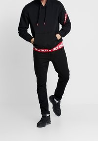 Felpa nera con cappuccio e tasca frontale, abbinata a jeans neri con una cintura rossa "REMOVE BEFORE" e sneaker neri.