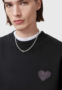 Felpa nera con scollatura a costine, caratterizzata da un piccolo design ricamato a forma di cuore in grigio. Indossata sopra un sottogiacca bianco con catena argentata.