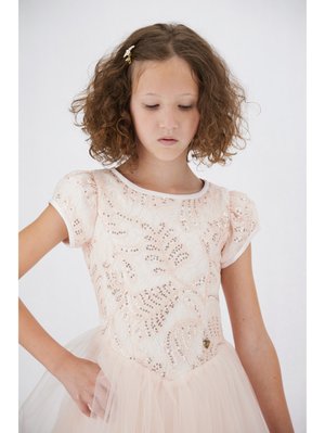 Młoda dziewczyna o kręconych włosach, ubrana w jasnoróżową sukienkę z cekinowymi kwiatowymi wzorami i tiulową spódnicą, patrząca w dół z zamyśleniem.