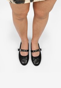Chaussures Mary Jane noires en cuir avec un bout rond, dotées d'un nœud décoratif, d'une bride de cheville et d'une boucle argentée avec des clous.
