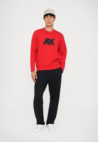 Červená mikina s čiernym logom "AX", v kombinácii s čiernymi teplákmi a bielymi teniskami. Model má na sebe svetlošedú čiapku. Jednoduché pozadie.