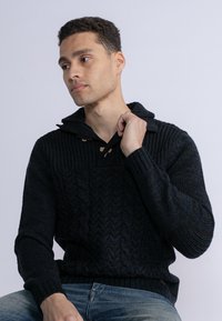 Pulover negru din tricot cu glugă, având un design texturat și mânere din lemn, asortat cu blugi albaștri.