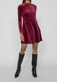 Mörkröd sammet långärmad klänning med en figurnära överdel och kjol som vidgar sig; kombinerad med svarta knähöga spetsiga stövlar. Slät textur genom hela plagget.