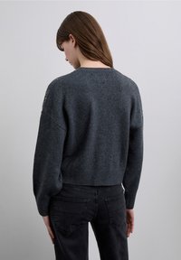 Maglione grigio lavorato a maglia con scollo rotondo, maniche a palloncino e orlo a coste; abbinato a jeans neri, mostrando una texture morbida.