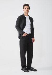 Cardigan noir zippé sur un t-shirt blanc, associé à un pantalon noir et des chaussures noires. Design simple avec une texture lisse et aucun motif visible.