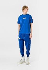 T-shirt de algodón azul con mangas cortas y bolsillo frontal. Los pantalones de chándal azules a juego cuentan con cordón lateral, acento blanco y estampado gráfico.