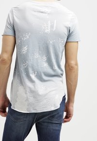 Camiseta azul claro con un patrón blanco salpicado, escote redondo, mangas cortas y un dobladillo trasero ligeramente alargado. Llevada con vaqueros oscuros.