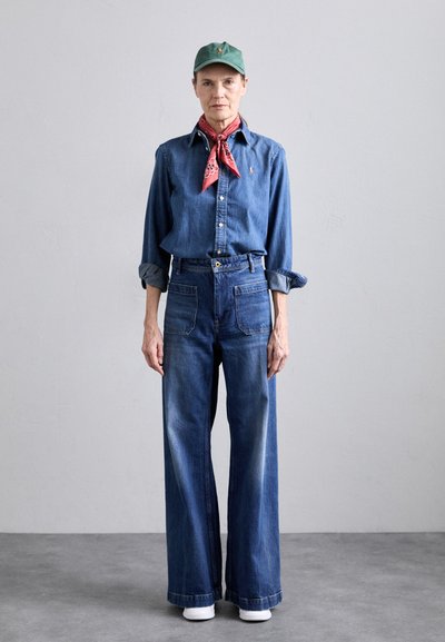 Denim shirt and wide-leg jeans in dark blue. White sneakers, green cap, and red bandana add contrast. Fabric appears sturdy with a smooth finish.