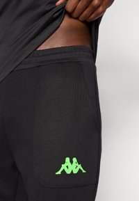 Čierne športové nohavice s jemnou textúrou, s predným vreckom, elastickým pásom a zeleným logom Kappa na boku.