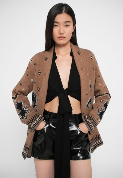 Cardigan en tricot marron avec des motifs géométriques noir et blanc, à l'avant ouvert et à manches longues, associé à un short noir brillant.
