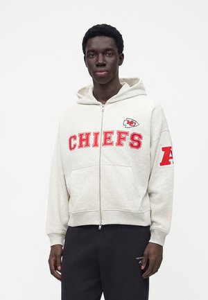 Jeune homme portant un sweat à capuche gris clair zippé avec "CHIEFS" et un logo rouge sur la poitrine ainsi qu'un lettrage rouge sur la manche.