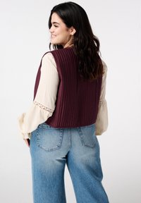 Bordeauxrood geribbeld hemd over een crèmekleurige blouse met kanten accenten, gecombineerd met lichtblauwe wijde jeans. De stof lijkt textuurvol en gelaagd.
