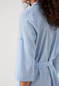 Camisa riscada azul claro com mangas de três quartos, detalhe de botão na manga e um cinto à volta da cintura. Tecido suave e texturizado.