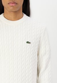 Hvit kabelstrikket genser med rund hals. Har en liten grønn krokodille-logo på brystet. Myk tekstur med ribbestruktur.