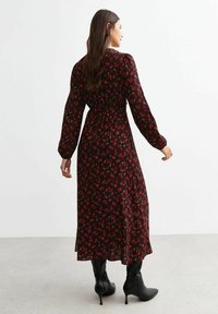 Robe florale noire avec un motif de fleurs rouges, manches longues, taille cintrée et jupe fluide. Le modèle porte des bottes noires montantes.