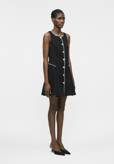 Γυναίκα που στέκεται φορώντας ένα μαύρο αμάνικο φόρεμα με λευκά κουμπιά στο μπροστινό μέρος και ταιριαστά μαύρα παπούτσια με μυτερή μύτη.