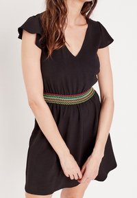 Schwarzes Kleid mit kurzen Rüschensäumen und V-Ausschnitt. Verfügt über einen mehrfarbigen, gewebten Gürtel mit roten, grünen und gelben Akzenten.