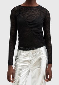 Personne portant un haut noir à manches longues en dentelle avec un design froncé et un pantalon métallique argenté brillant sur un fond uni.