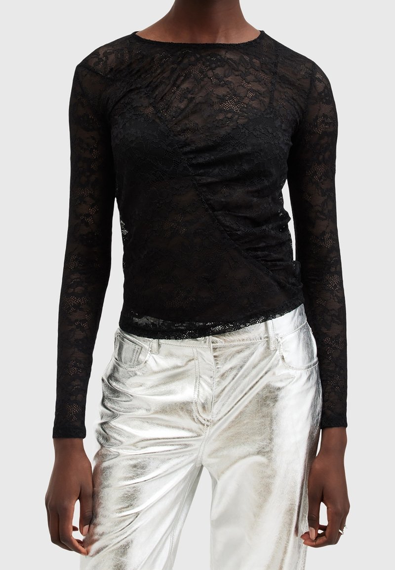 Personne portant un haut noir à manches longues en dentelle avec un design froncé et un pantalon métallique argenté brillant sur un fond uni.