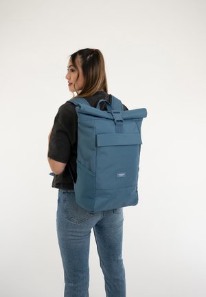 Blue roll-top backpack made of durable fabric, featuring a front pocket, adjustable straps, and a logo patch at the bottom.