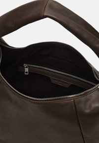 Wnętrze brązowej skórzanej torby z kieszenią na zamek, gładką teksturą, zaokrąglonym uchwytem oraz charakterystyczną metką. Widoczna ciemna podszewka materiałowa.