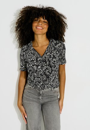 Femme aux cheveux bouclés souriante, vêtue d'un chemisier à manches courtes noir et blanc à motifs floraux et d'un jean gris taille haute sur fond blanc.