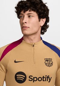 Beige urheilupaita pitkillä hihoilla, jossa on neljännesvetoketju, musta Nike-logo, Barcelona-merkki ja moniväriset olkaviivat sinisessä ja viininpunaisessa.