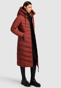 Long, rust-colored puffer coat with a hood, quilted design, two side pockets, front zipper, and accent drawstring details. Textured finish.