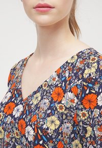 Haut en tissu à motif floral avec un décolleté en V profond. Les couleurs incluent marine, orange, jaune et blanc. Texture légère et douce.