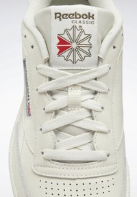 Nära bild av vita Reebok Classic-sneakers med detaljerad logotyp, röd och brun stjärndesign, och prydligt knutna vita skosnören.