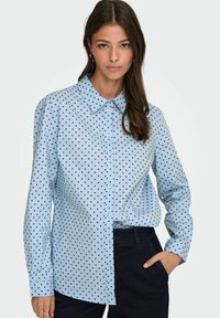 Chemise boutonnée bleu clair avec des pois navy, col classique, manches longues et coupe décontractée ; le tissu semble lisse et léger.