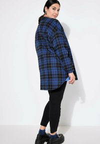 Camicia oversize a quadri neri e blu con maniche lunghe, spalle cadenti, fessure laterali e orlo curvo, abbinata a leggings neri e scarpe chunky.