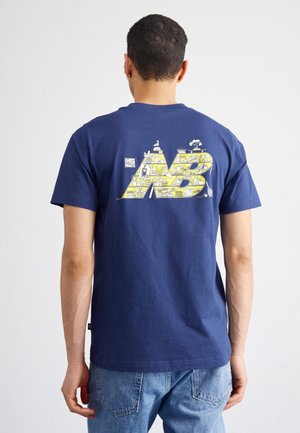 Mann trägt ein marineblaues T-Shirt mit großem gelbem "NB"-Logo, das auf der Rückseite mit kleinen weißen Illustrationen gefüllt ist, kombiniert mit blauen Jeans.
