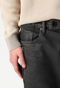 Vaqueros de denim negro con una textura desgastada, que cuentan con un zíper en el bolsillo lateral y un cierre de botón. Combinados con un suéter de canalé en color beige claro.