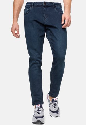 Jeans oscuros de corte ajustado con cinco bolsillos, un ligero desgaste y un material de denim ligero, combinados con zapatillas grises y azul marino.
