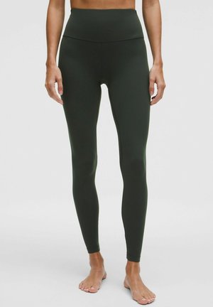 Osoba oblečená v tmavozelených legínach s vysokým pásom, stojaca naboso na svetlom jednom pozadí.