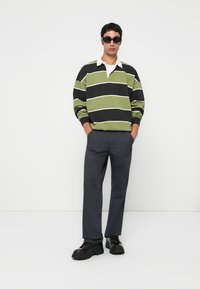Polo a maniche lunghe a righe verdi e nere con dettagli bianchi, abbinato a pantaloni scuri e scarpe nere chunky. Il modello sta in piedi con le mani in tasca.
