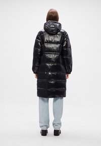 Melna pūkaina jaka ar spīdīgu, pildītu tekstūru, kapuci un garām piedurknēm, kas sasniedz vidus teļa; apvienota ar gaiši ziliem džinsiem un masīviem melniem zābakiem.