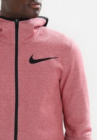 Rote Fleece-Hoodie mit hohem Kragen und schwarzem Reißverschluss. Hat ein auffälliges schwarzes Nike-Logo auf der linken Brust. Glatte Textur, athletischer Schnitt.