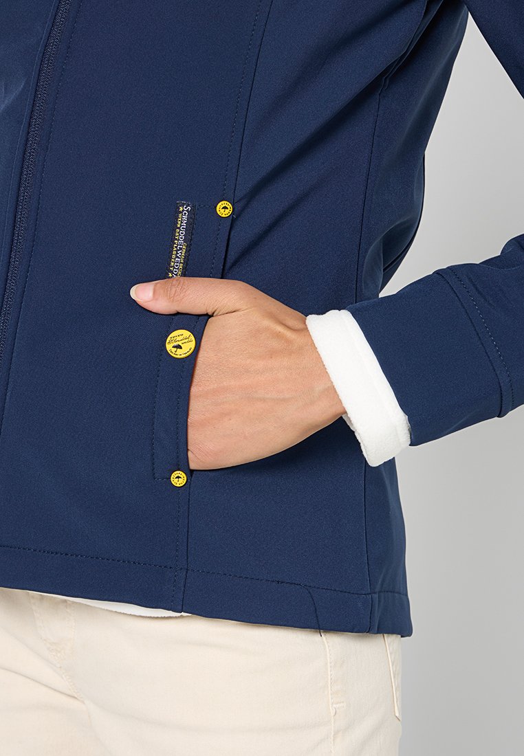 Dunkelblaue Softshelljacke mit einer Reißverschlusstasche, gelben Knopfakzenten und einem weißen, fleecegefütterten Saum. Nahaufnahme der Hand in der Tasche.